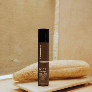 Commen Nº.11 Anti-Age Day Cream on a stone tray in front of a loofah, in a warm bathroom.