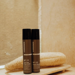 Commen Nº.12 Matte Zinc Gel next to Nº.11 Anti-Age Day Cream on a stone tray with a loofah, in a warm bathroom.