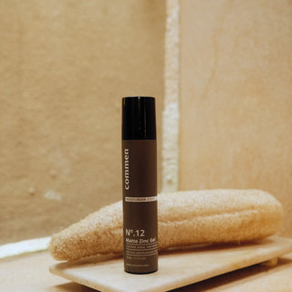 Commen Nº.12 Matte Zinc Gel on a stone tray in front of a loofah, in a warm bathroom.