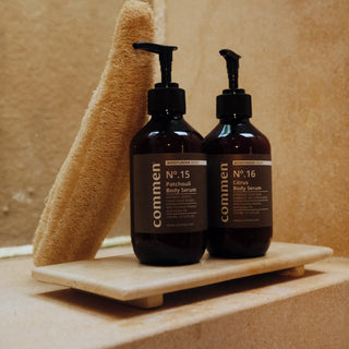 Commen Nº.16 Citrus Body Serum next to Nº.15 Patchouli Body Serum on a stone tray with a loofah, in a warm bathroom.