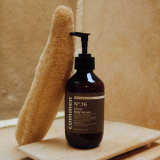 Commen Nº.16 Citrus Body Serum on a stone tray beside a loofah, in a warm bathroom.