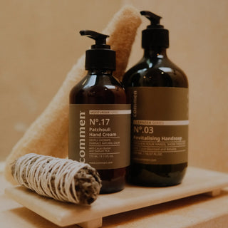 Commen Nº.17 Patchouli Hand Cream met Nº.03 Revitalising Handsoap op een stenen plankje, met een luffa en een bundel salie.