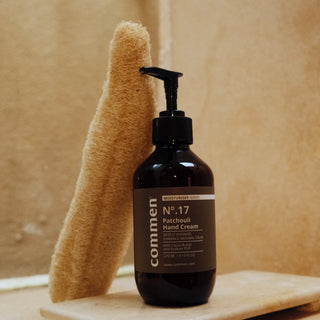Commen Nº.17 Patchouli Hand Cream op een stenen plankje naast een luffa, in een warme badkamer.