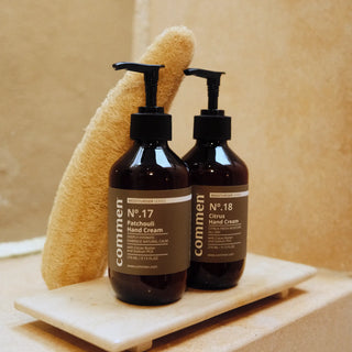 Commen Nº.18 Citrus Hand Cream next to Nº.17 Patchouli Hand Cream on a stone tray with a loofah, in a warm bathroom.