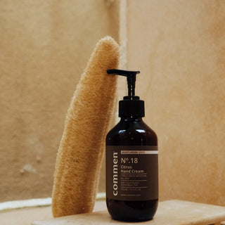 Commen Nº.18 Citrus Hand Cream on a stone tray beside a loofah, in a warm bathroom.
