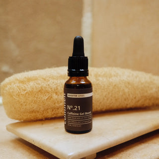 Commen Nº.21 Caffeine Gel Boost on a stone tray in front of a loofah, in a warm bathroom.