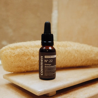 Commen Nº.22 Ginkgo Antioxidant Boost on a stone tray beside a loofah, in a sunlit bathroom.