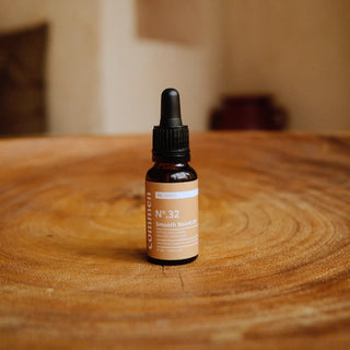 Commen Nº.32 Smooth Beard Oil, een amberkleurig pipetflesje op een ronde teakhouten tafel in een lichte gestucte kamer.