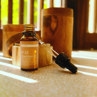 Commen Nº.32 Smooth Beard Oil, een open amberkleurige fles met de glazen pipet ernaast, in warm gestreept zonlicht.