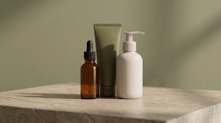 Three unbranded skincare containers, an amber dropper bottle, a sage tube and a cream pump bottle, standing together on a travertine ledge against an olive wall.